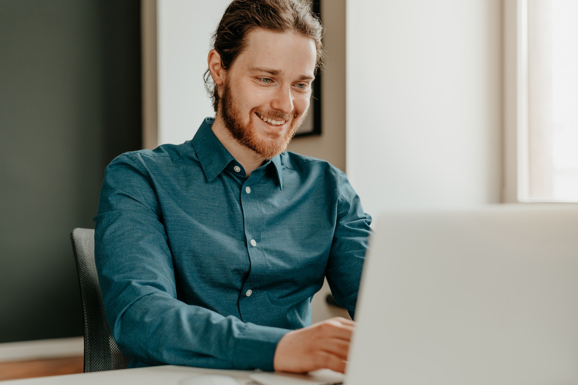 Smiling young male office worker typing on keyboard and looking at laptop screen in office.